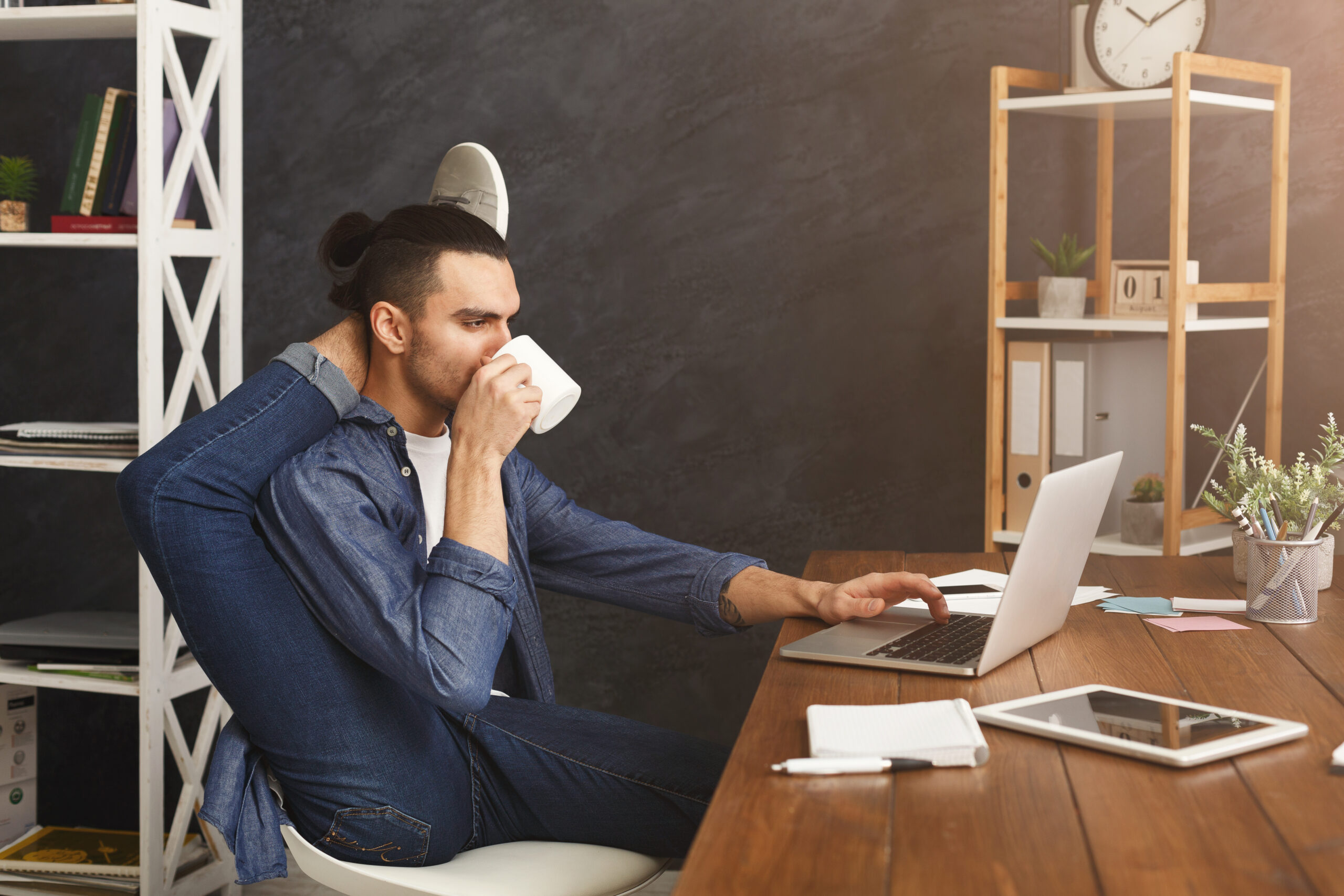 Short break for yoga in office. Flexible man practicing yoga at workplace, while typing on laptop and having coffee, copy space. Active employee at work, healthy lifestyle concept