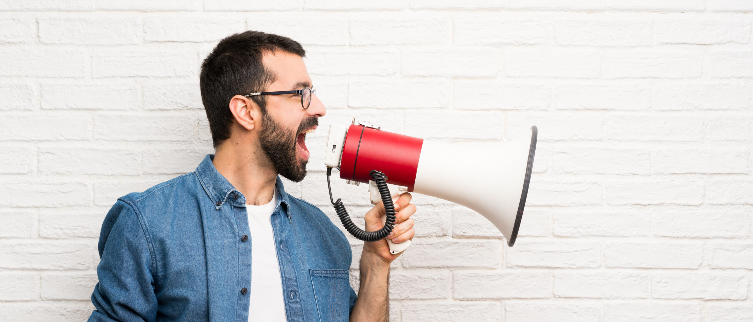 Handsome man with beard over white brick wall shouting through a megaphone