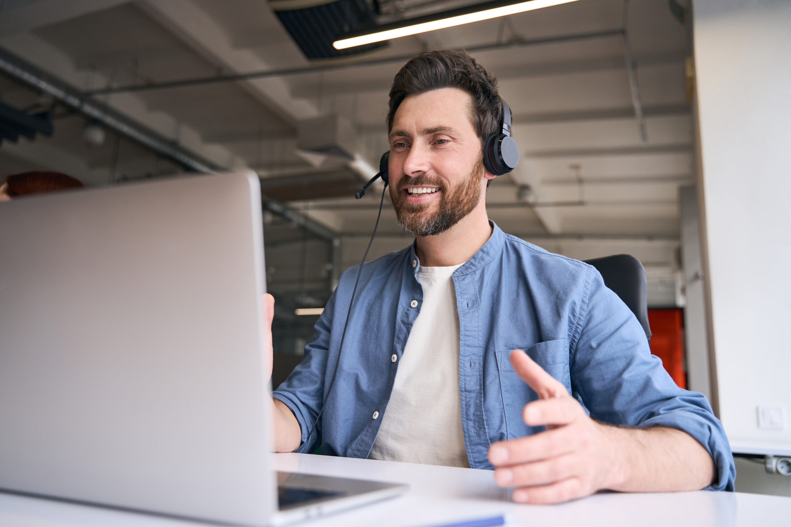 Handsome smiling man, call center operator using laptop, answering call, communication with customer sitting in modern office. Freelancer having video call at workplace